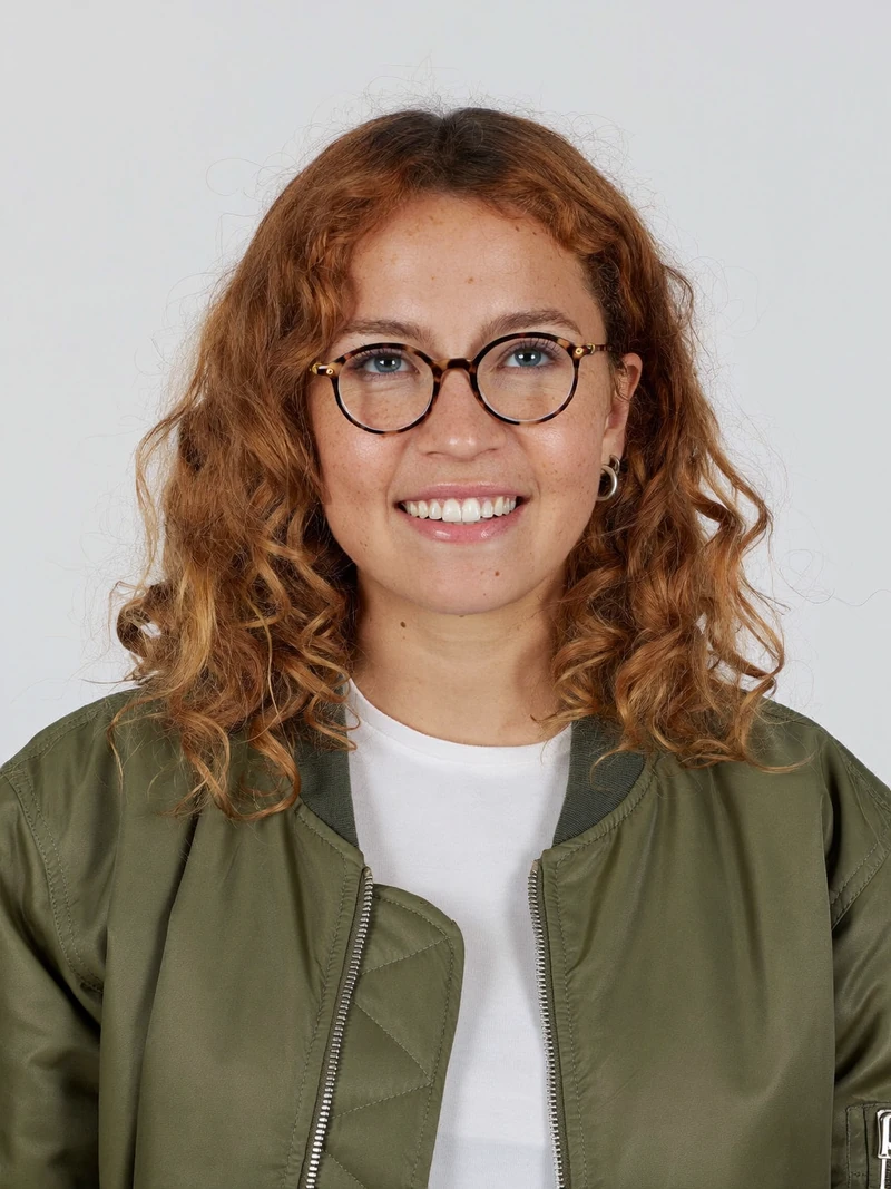AI-generated portrait of our fictional test character: a smiling woman with curly copper hair, round tortoiseshell glasses and an olive bomber jacket over a white t-shirt, light-gray background
