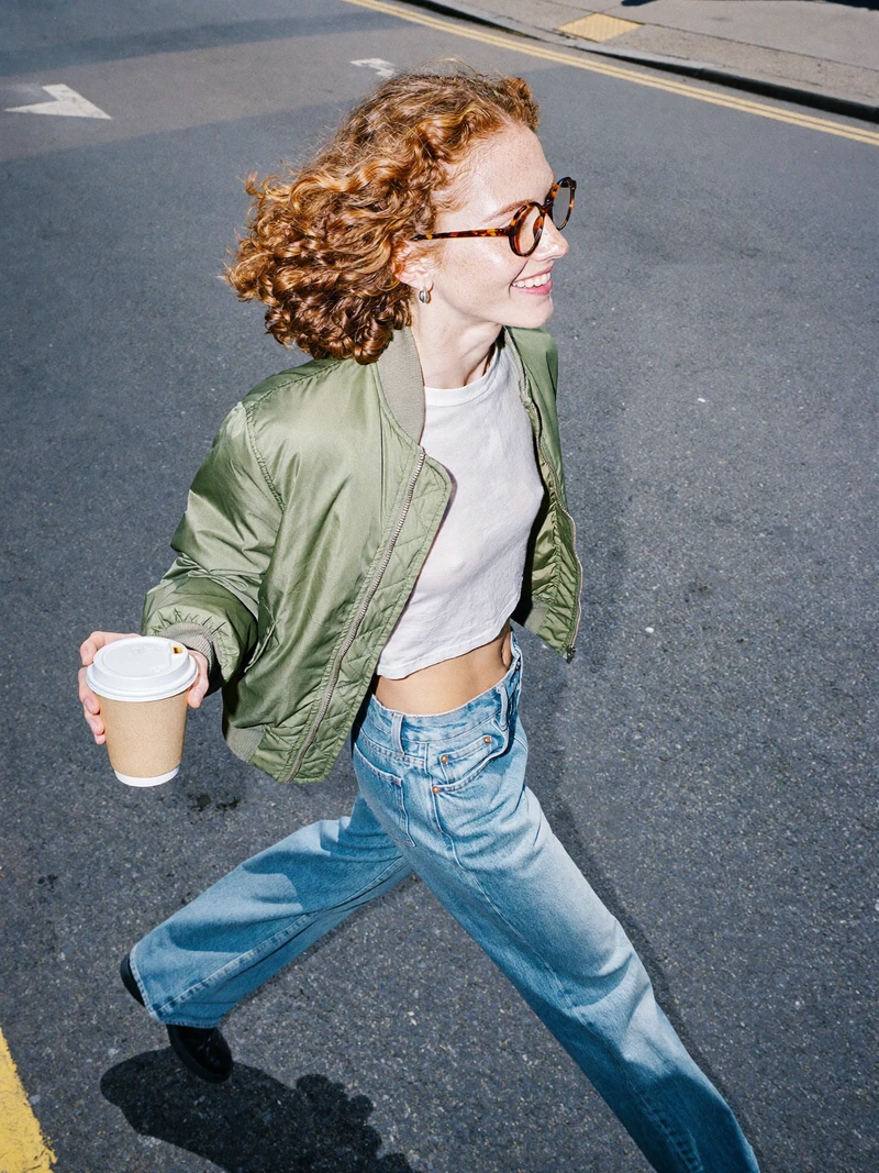 AI-generated photo of a fictional adult woman with copper curls and round glasses striding across a street, high angle