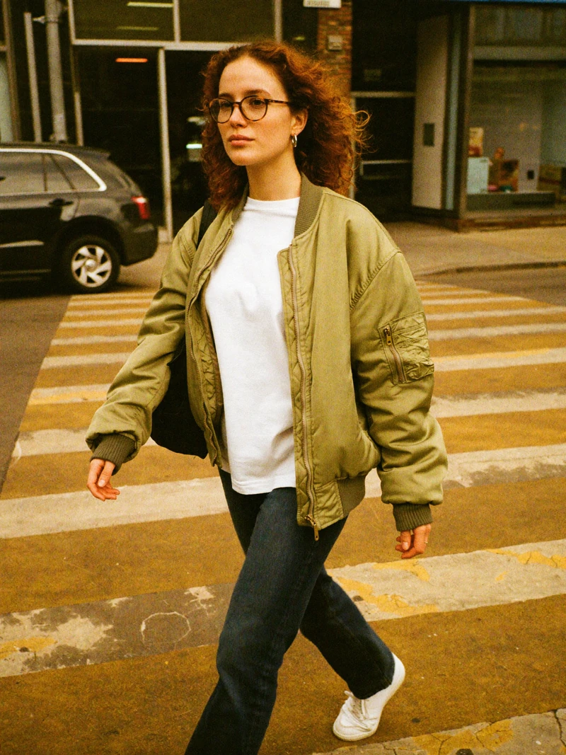 AI-generated photo of a fictional adult woman in an olive bomber jacket and jeans walking on a crosswalk, warm film color