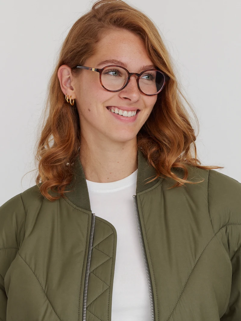 AI-generated waist-up photo of a fictional adult woman with wavy copper hair and round glasses on a gray studio backdrop