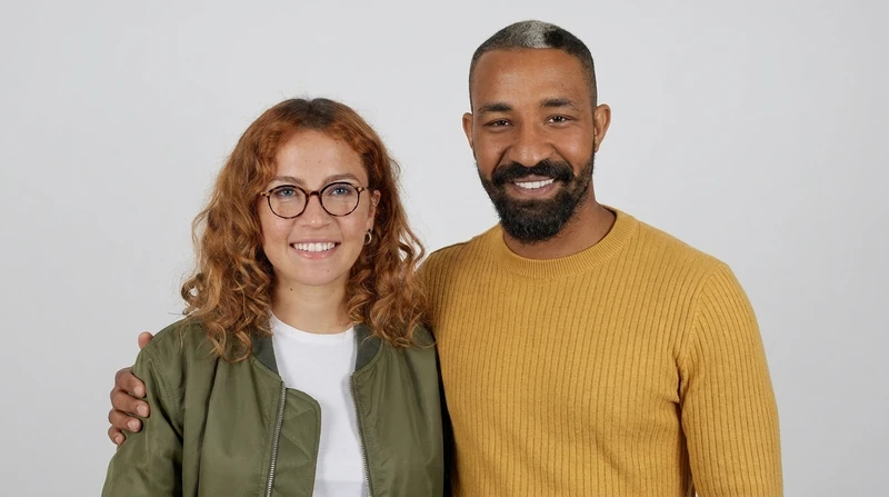 AI-generated start frame: a fictional woman with copper curls and round glasses and a fictional bearded man in a mustard sweater, side by side on a light-gray backdrop, his arm around her shoulders
