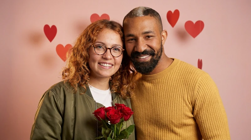 Two fictional adults in front of a blush-pink backdrop with red paper hearts, a bouquet of red roses in front of her, their heads tilted together, no kiss